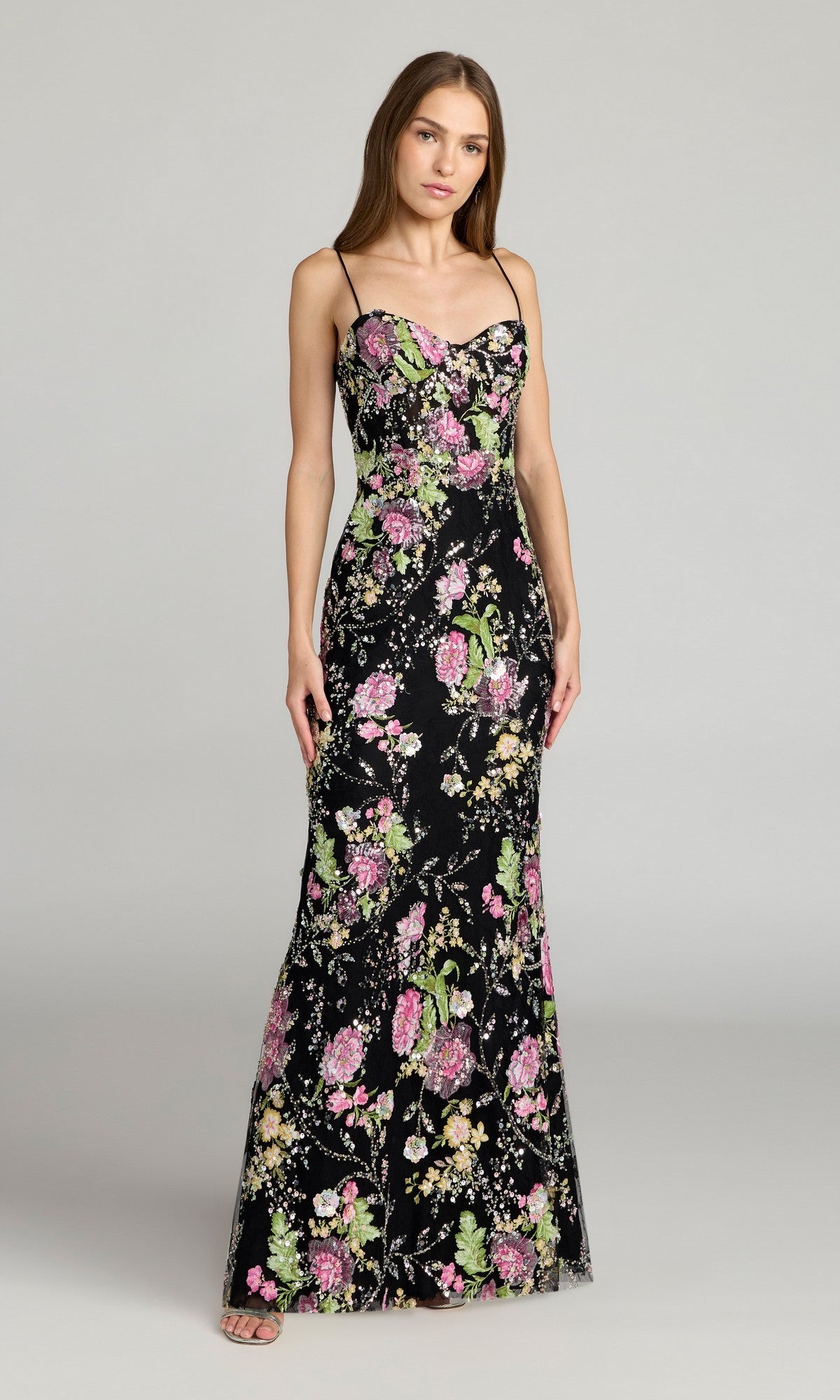 A woman wears the Mac Duggal 68891 Beaded Long Floral Formal Dress, a black, sleeveless gown with thin straps and vibrant floral detailing, standing against a plain gray background.